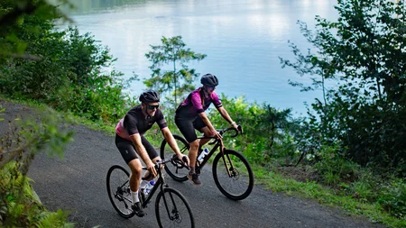 Zwei Radfahrer fahren auf einem Waldweg neben einem See bei bewölktem Himmel