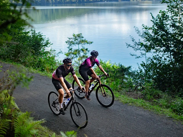 Zwei Radfahrer fahren auf einem Waldweg neben einem See bei bewölktem Himmel