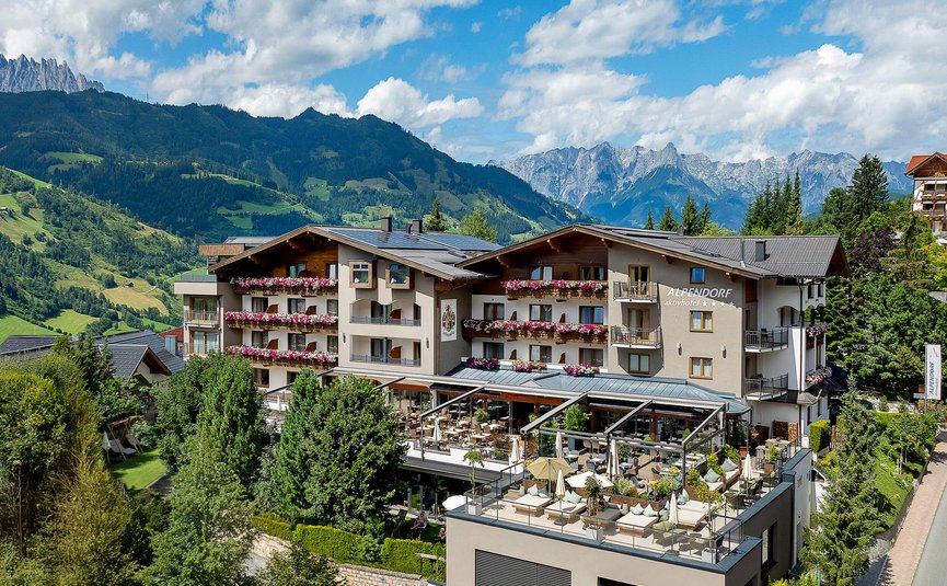 Hotel Alpendorf mit Terrasse in bergiger Landschaft bei Sonnenschein