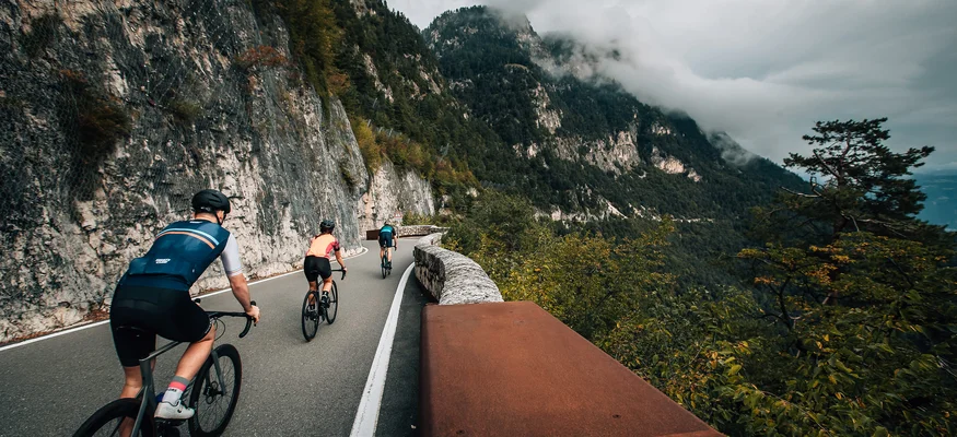 Three cyclists riding on mountain road with forested hills and clouds