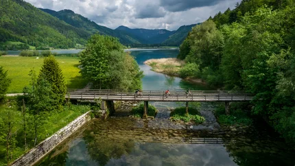 Cyclists on wooden bridge over river in green landscape with mountains and cloudy sky