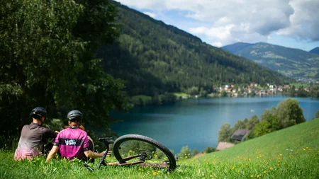 Zwei Radfahrer sitzen auf einer Wiese mit Blick auf See und Berge.