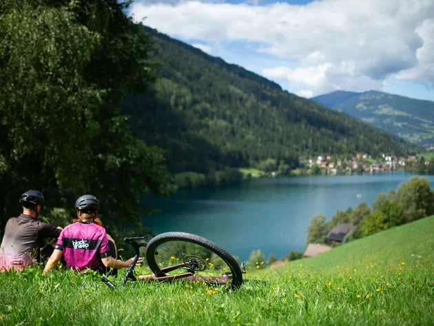 Zwei Radfahrer sitzen auf einer Wiese mit Blick auf See und Berge.