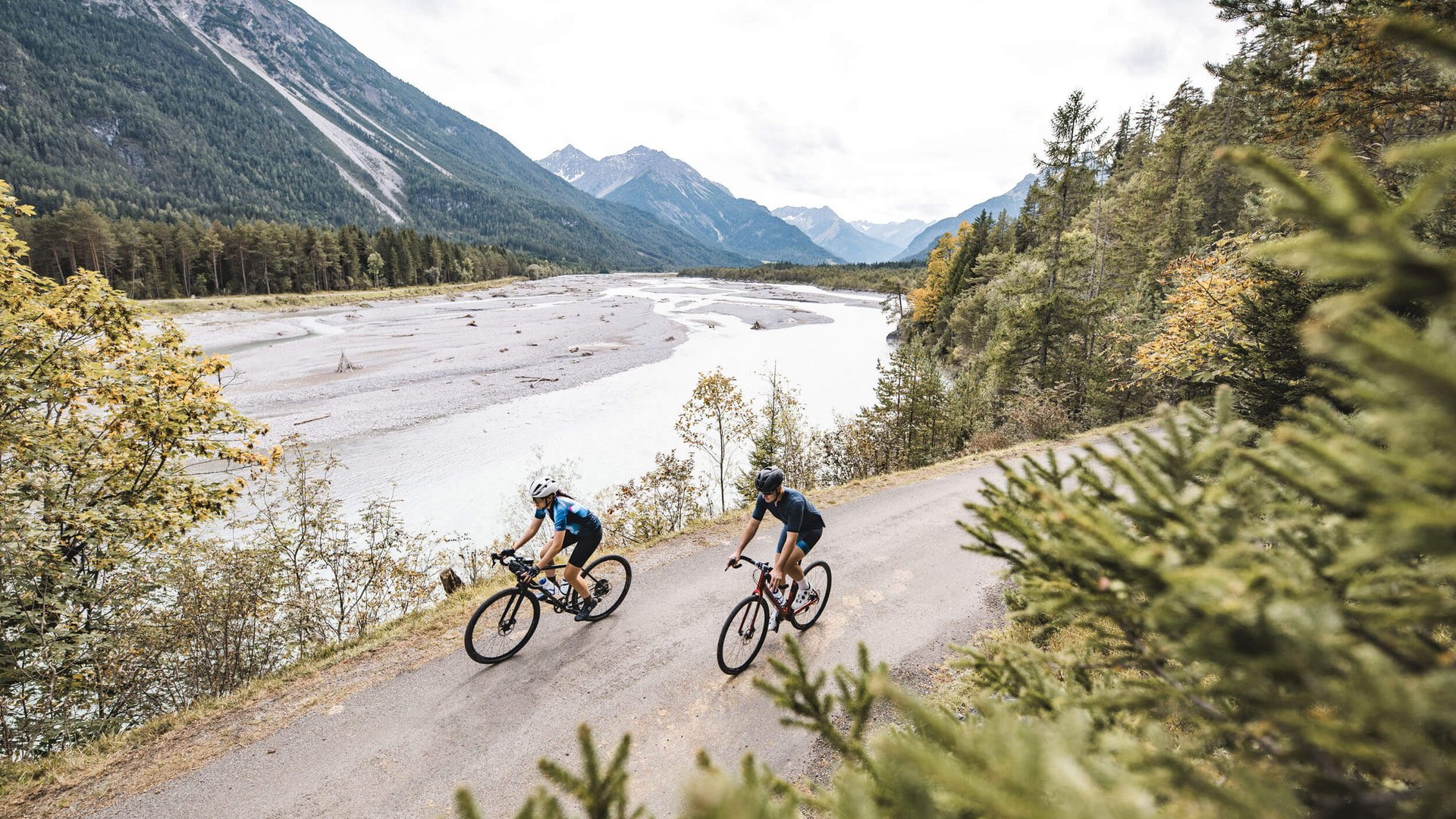 Zwei Radfahrer fahren auf einem Bergweg neben einem Fluss mit Bergen im Hintergrund