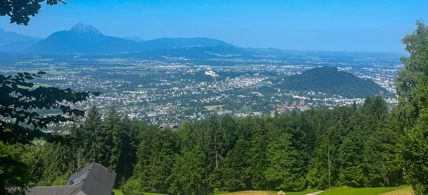 Blick auf Stadt und Berge unter blauem Himmel von einem Hügel mit Bäumen und Wiesen.