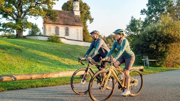 5 Tipps fürs Graveln in Oberbayern © Oberbayern.de Zwei Radfahrer vor einer kleinen Kirche auf Landstraße im Grünen
