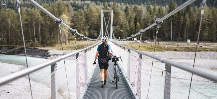 Person schiebt Fahrrad auf Hängebrücke über Fluss in bewaldeter Berglandschaft