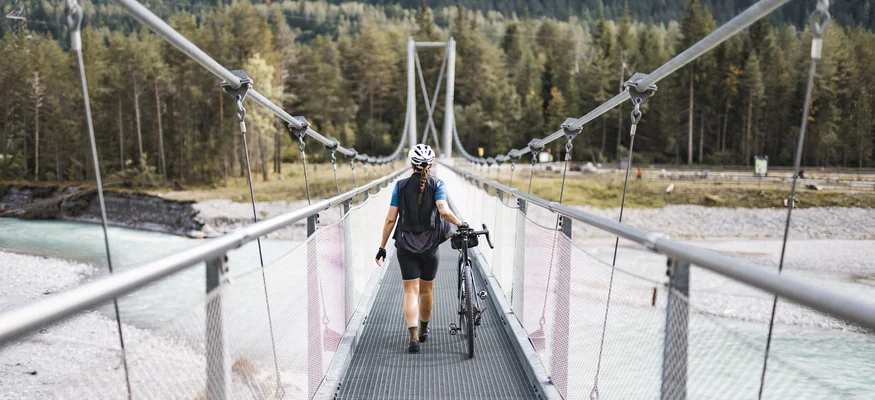 Person schiebt Fahrrad auf Hängebrücke über Fluss in bewaldeter Berglandschaft