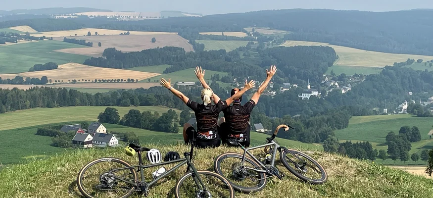 Zwei Radfahrer sitzen mit erhobenen Armen auf einem Hügel mit Blick auf das Tal