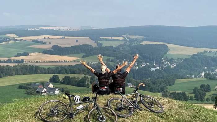 Two cyclists sitting on a hill with raised arms overlooking a valley