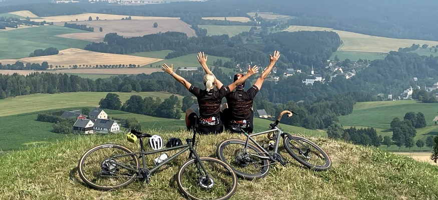 Zwei Radfahrer sitzen mit erhobenen Armen auf einem Hügel mit Blick auf das Tal
