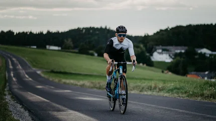 Radfahrer auf Landstraße mit grüner Wiese und Häusern im Hintergrund
