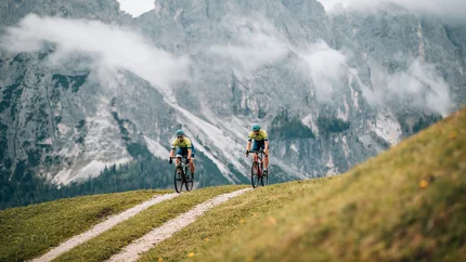 Two cyclists riding on a mountain trail with misty alpine cliffs