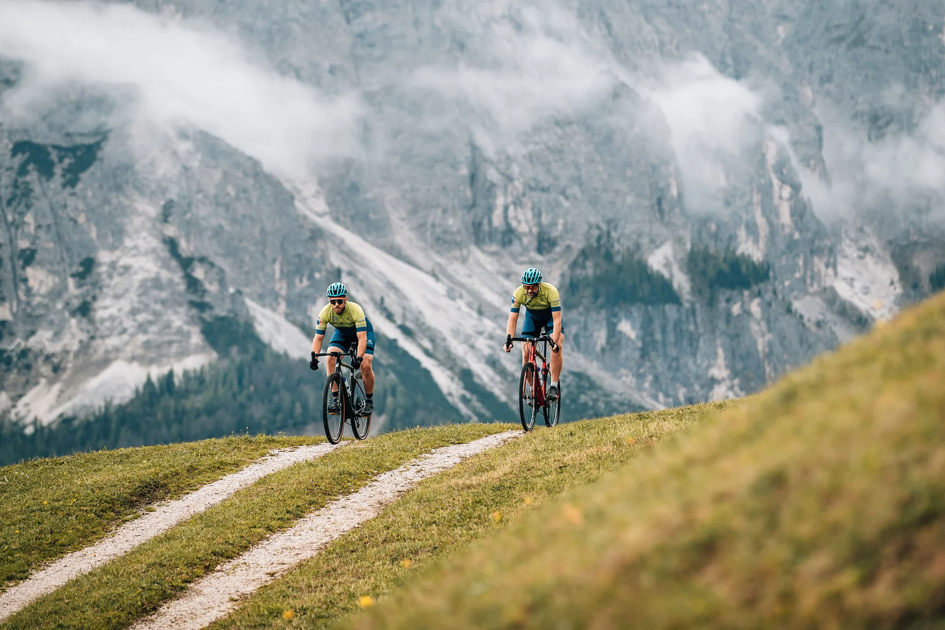 Gravelbike Holidays © Jens Scheibe Zwei Radfahrer fahren auf einem Bergweg vor nebligen Alpenfelsen