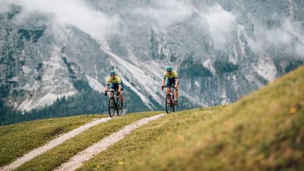 Two cyclists riding on a mountain trail with misty alpine cliffs