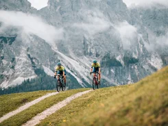 Kronplatz Dolomites region © Jens Scheibe Two cyclists riding on a mountain trail with misty alpine cliffs