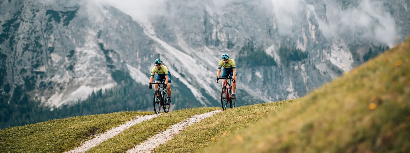 Juvel & Armentara © Jens Scheibe Two cyclists riding on a mountain trail with misty alpine cliffs