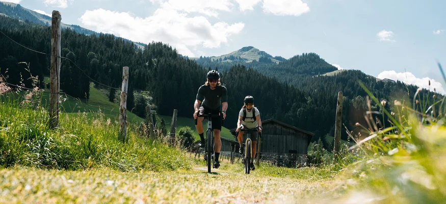 Graveln in den Kitzbüheler Alpen © Martina Wunderle / Flo Distl Zwei Radfahrer fahren auf einem Weg in den Bergen bei sonnigem Wetter