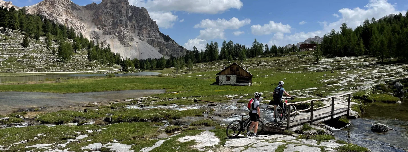 Passo Furcia round © Hotel Ostaria Posta Two cyclists crossing a bridge in a mountain landscape with a cabin