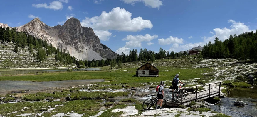 Zwei Radfahrer überqueren eine Brücke in einer alpinen Berglandschaft mit Hütte
