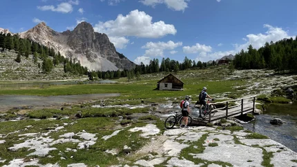 Zwei Radfahrer überqueren eine Brücke in einer alpinen Berglandschaft mit Hütte