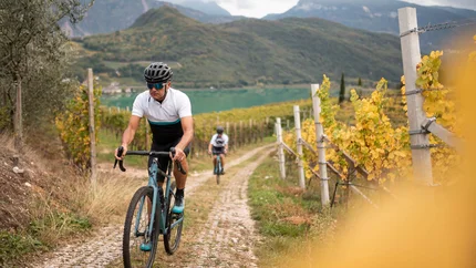 Cyclists riding on a gravel path between vineyards with mountains behind