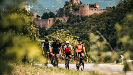 Gravel & Törggelen: Warum Tramin im Herbst perfekt für Gravelbiker ist © Jens Scheibe Vier Radfahrer fahren auf einer Straße vor einer alten Burg in einer grünen Landschaft