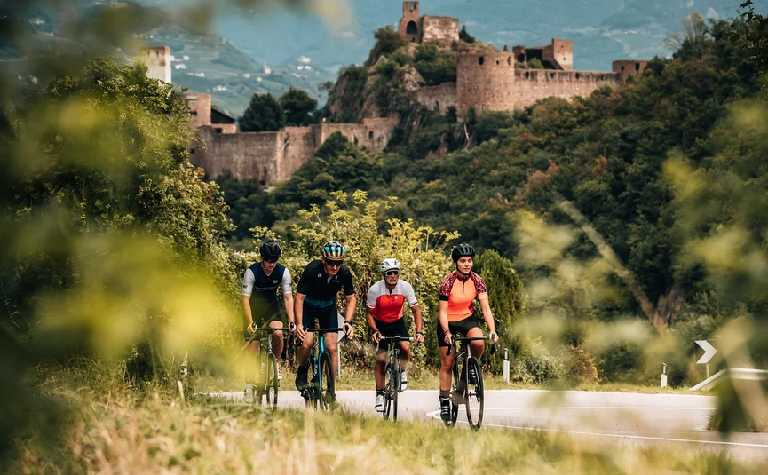 Vier Radfahrer fahren auf einer Straße vor einer alten Burg in einer grünen Landschaft
