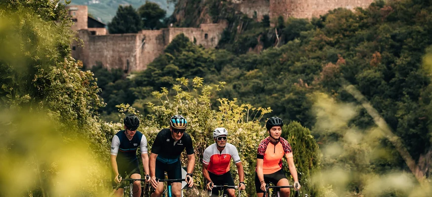 Genuss & Gravel in Tramin © Jens Scheibe Vier Radfahrer fahren auf einer Straße vor einer alten Burg in einer grünen Landschaft