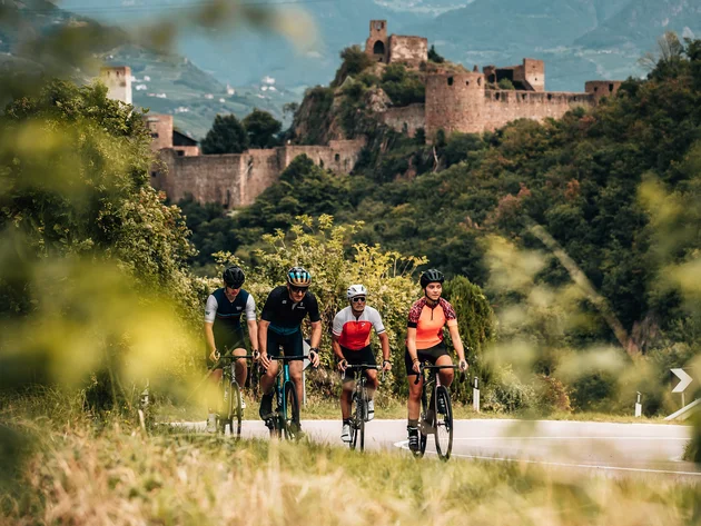Gravel & Törggelen: Warum Tramin im Herbst perfekt für Gravelbiker ist © Jens Scheibe Vier Radfahrer fahren auf einer Straße vor einer alten Burg in einer grünen Landschaft
