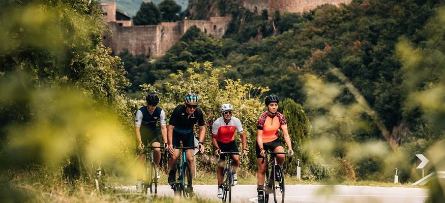 Vier Radfahrer fahren auf einer Straße vor einer alten Burg in einer grünen Landschaft