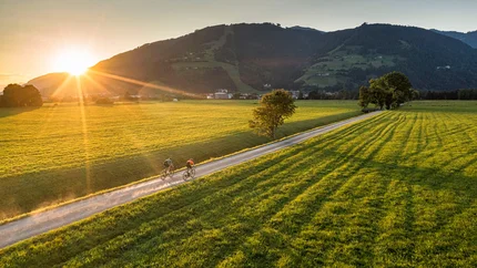 5 Gravel-Touren in der Salzburger Sportwelt, die du kennen solltest © SalzburgerLand Tourismus Zwei Radfahrer auf Landweg bei Sonnenuntergang in grüner Berglandschaft
