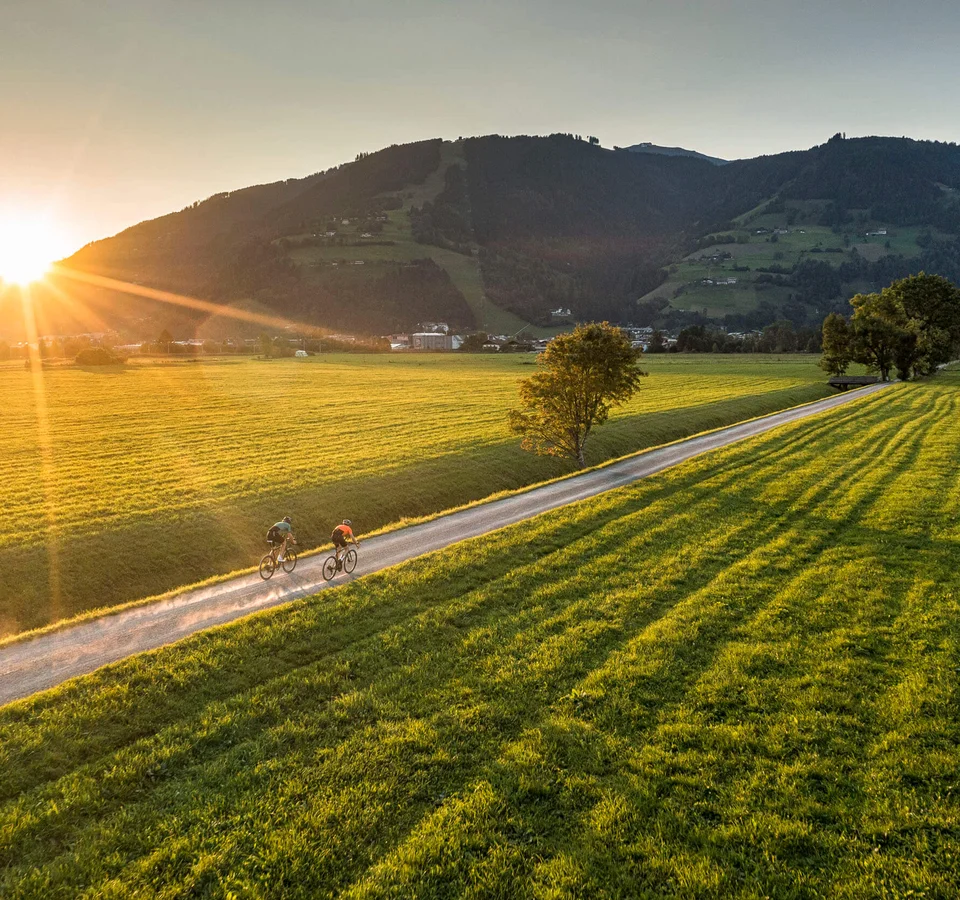 5 Gravel-Touren in der Salzburger Sportwelt, die du kennen solltest © SalzburgerLand Tourismus Zwei Radfahrer auf Landweg bei Sonnenuntergang in grüner Berglandschaft