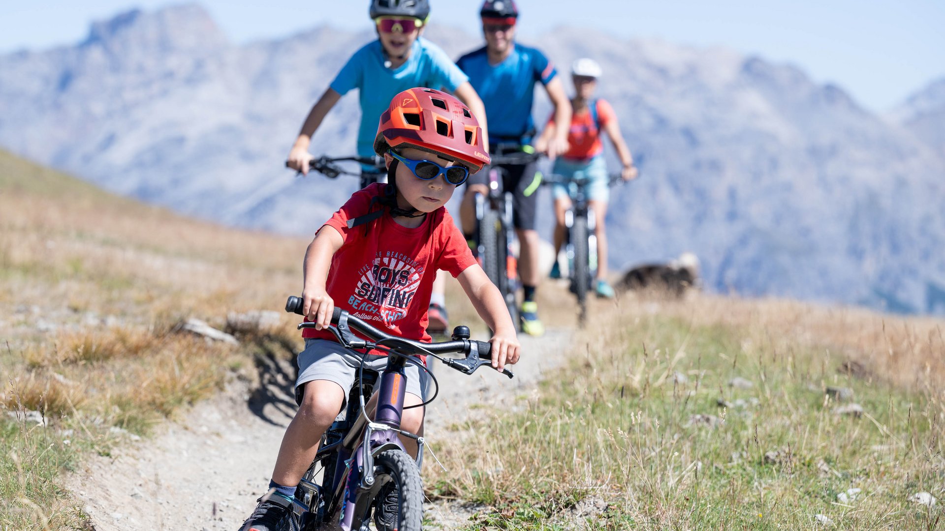 Kind mit rotem Helm fährt Fahrrad bergab, Familie folgt auf Mountainbikes