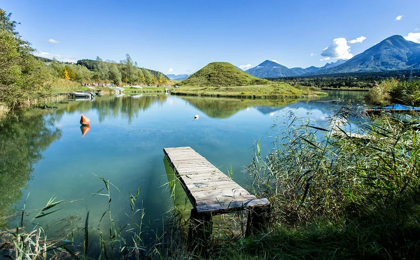 Lake with wooden pier, green hills, and mountains in the background