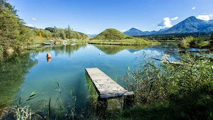 Lake with wooden pier, green hills, and mountains in the background