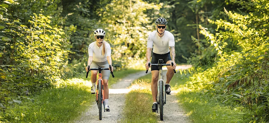 Zwei Radfahrer in weißer Sportkleidung fahren auf einem Waldweg