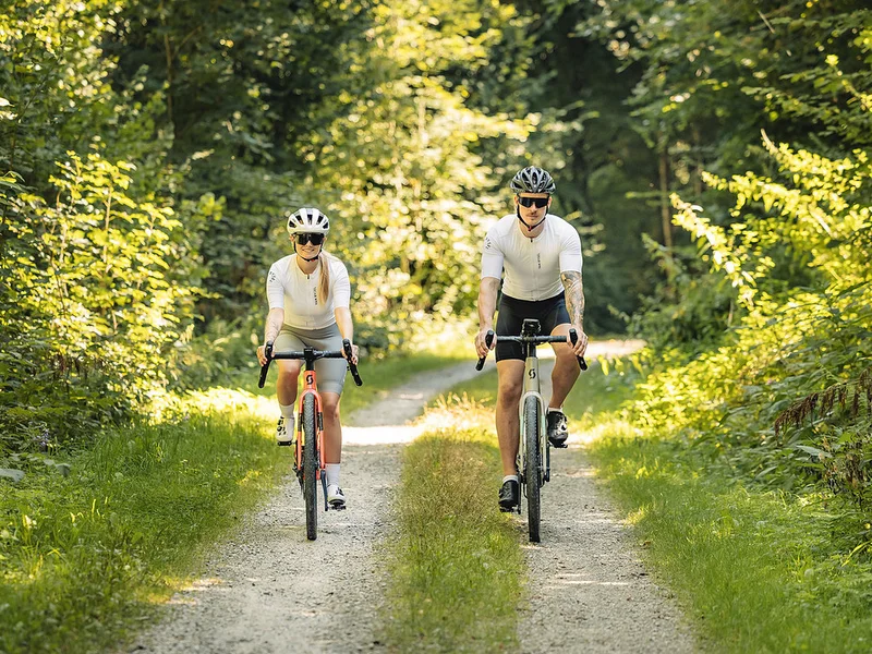 Gravelbike Holidays © Stefan J. Wolf Photography Zwei Radfahrer in weißer Sportkleidung fahren auf einem Waldweg
