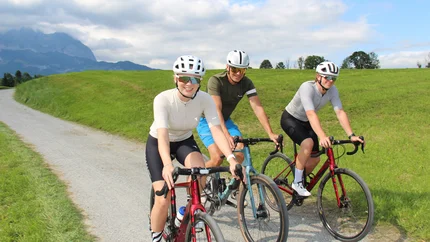 Drei Radfahrer auf einem Landweg mit grünen Wiesen und Bergen im Hintergrund